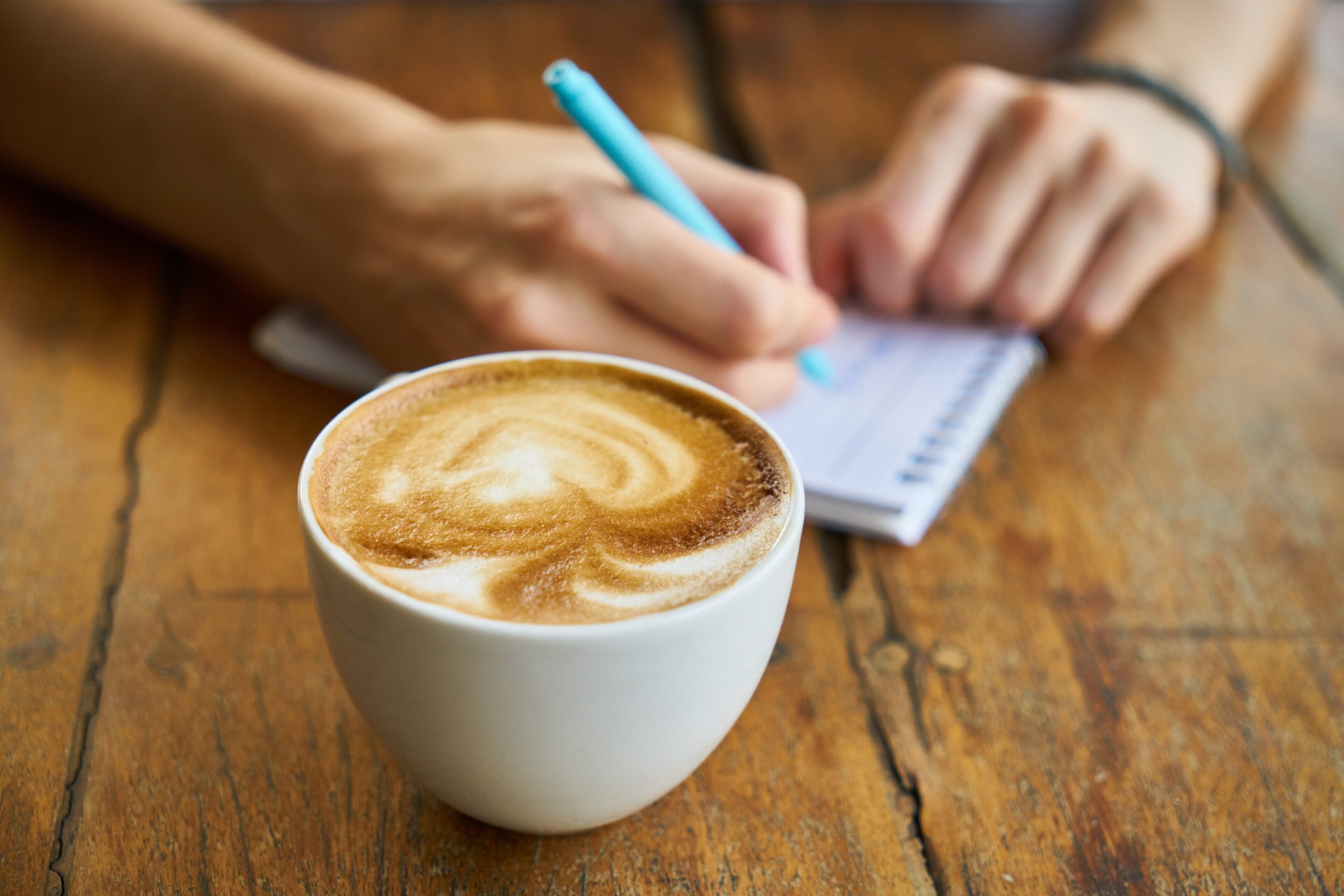 A cup of cappuccino with latte art on a wooden table and a person writing notes.